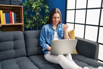 Wall Mural - Hispanic woman using laptop at home annoyed and frustrated shouting with anger, yelling crazy with anger and hand raised