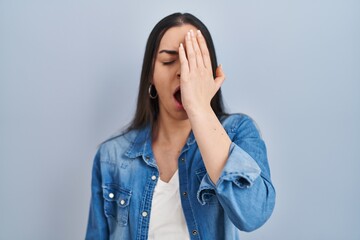 Wall Mural - Hispanic woman standing over blue background yawning tired covering half face, eye and mouth with hand. face hurts in pain.