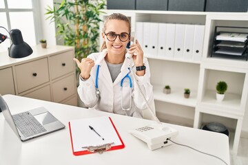 Canvas Print - Young doctor woman speaking on the phone pointing thumb up to the side smiling happy with open mouth