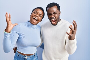 Poster - Young african american couple standing over blue background crazy and mad shouting and yelling with aggressive expression and arms raised. frustration concept.