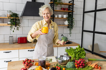 Wall Mural - Attractive senior woman with grey hair cooking healthy food on her kitchen at home. Mature female preparing fresh vegetarian salad with organic ingredients from the market: tomato, cucumber, pepper