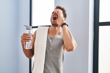 Poster - Young hispanic man wearing sportswear drinking water shouting and screaming loud to side with hand on mouth. communication concept.