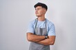 Hispanic young man wearing apron over white background looking to the side with arms crossed convinced and confident