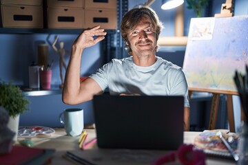 Poster - Middle age man sitting at art studio with laptop at night gesturing with hands showing big and large size sign, measure symbol. smiling looking at the camera. measuring concept.