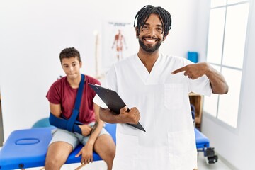 Wall Mural - Young hispanic man working at pain recovery clinic with a man with broken arm looking confident with smile on face, pointing oneself with fingers proud and happy.