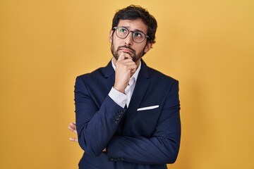 Sticker - Handsome latin man standing over yellow background with hand on chin thinking about question, pensive expression. smiling with thoughtful face. doubt concept.