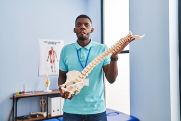 Wall Mural - African american man holding anatomical model of spinal column making fish face with mouth and squinting eyes, crazy and comical.