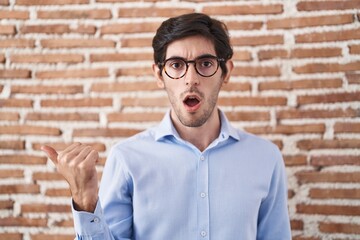 Canvas Print - Young hispanic man standing over brick wall background surprised pointing with hand finger to the side, open mouth amazed expression.