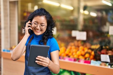 Young chinese woman employee talking on the smartphone using touchpad at fruit store