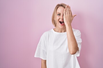 Poster - Young caucasian woman standing over pink background covering one eye with hand, confident smile on face and surprise emotion.