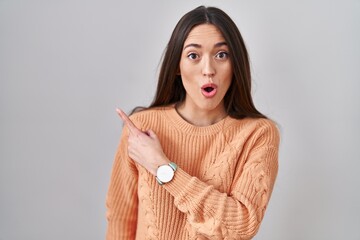 Wall Mural - Young brunette woman standing over white background surprised pointing with finger to the side, open mouth amazed expression.