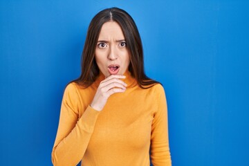 Wall Mural - Young brunette woman standing over blue background looking fascinated with disbelief, surprise and amazed expression with hands on chin