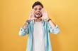 Young hispanic man with tattoos standing over yellow background smiling cheerful playing peek a boo with hands showing face. surprised and exited