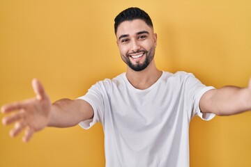 Poster - Young handsome man wearing casual t shirt over yellow background looking at the camera smiling with open arms for hug. cheerful expression embracing happiness.