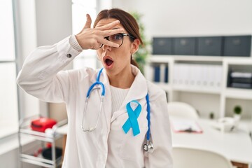 Canvas Print - Young brunette doctor woman wearing stethoscope at the clinic peeking in shock covering face and eyes with hand, looking through fingers with embarrassed expression.