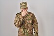 Young arab man wearing camouflage army uniform smelling something stinky and disgusting, intolerable smell, holding breath with fingers on nose. bad smell
