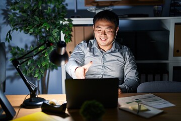 Canvas Print - Young chinese man working using computer laptop at night smiling friendly offering handshake as greeting and welcoming. successful business.