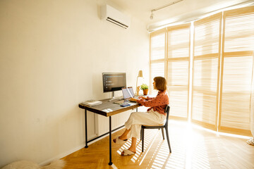 Wall Mural - Woman works on computers while sitting by a cozy workplace at sunny living room at home. Concept of remote work from home office