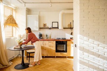 Woman puts flowers on a dinner table, decoration and having some house chores at home. Sunny interior of studio apartment in beige tones