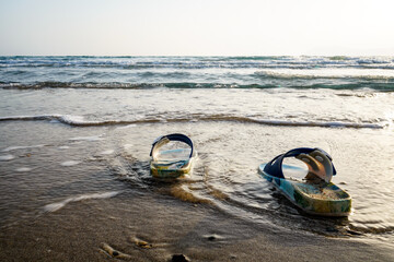 Wall Mural - focused colorful and sandy kids slippers sandals is on the beach, the sea and beach is blurred in the background, sunset and wavy sea