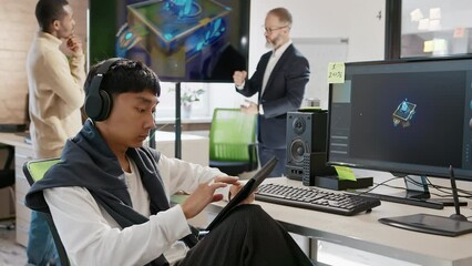Asian male artist works in headphones in a studio for the development of computer games and applications. In the background, a team of developers are discussing a 3D model.