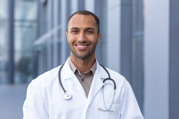 Portrait of successful and smiling African American doctor, man in medical coat with stethoscope looking at camera and smiling, outside modern hospital.