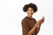 Portrait of young modern guy using mobile phone, looking behind his shoulder and smiling, standing with smartphone against white background
