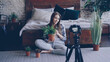 © silverkblack - Young smiling woman blogger in casual clothing is holding flowers, talking and recording video blog for online vlog about house plants using camera on tripod.
