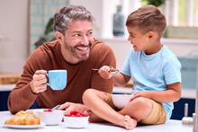 Family With Father In Kitchen With Son Sitting On Counter Eating Breakfast Together