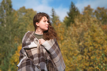 Wall Mural - portrait of beautiful cozy girl, young woman standing in golden autumn park or forest covering herself in blanket, plaid looking to the side. Copy space, place for text.