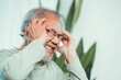 © Kiattisak - Closeup of Asian Senior elderly old man feel headache suffering and migraine in living room at home. Stressed mature male hurt mental health care and insurance concept.