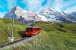 A cogwheel train travels on the railway from Jungfraujoch (top of Europe) to Kleine Scheidegg on the green grassy hillside with Eiger, Monch & Jungfrau in background in Bernese Highlands, Switzerland