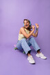 © Look! - Positive young caucasian girl smiling and sitting on skateboard on purple background in studio. Blonde wears headphones, top and jeans. Leisure, relaxation and lifestyle concept.