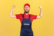 Portrait of proud strong worker man wearing blue uniform and red cap standing with raised arms and showing his power, looking at camera with pride. Indoor studio shot isolated on yellow background.