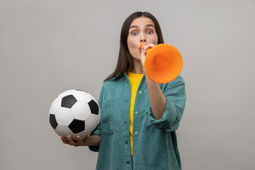 Portrait of excited woman blowing in horn holding black and white soccer ball, celebrating victory of favourite football team, wearing casual jacket. Indoor studio shot isolated on gray background.