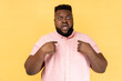 Portrait of bearded astonished man wearing pink shirt pointing at himself, asks who me, has surprised expression, shocked being picked. Indoor studio shot isolated on yellow background.