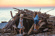 two little cheerful girls sisters of different ages are sitting in a homemade hut made of logs and sticks of trees on the seashore