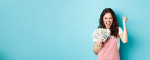 Lucky Young Woman Looks Excited, Shouting From Satisfaction And Triumph, Winning Money, Holding Dollar Bills And Making Fist Pump, Standing Over Blue Background