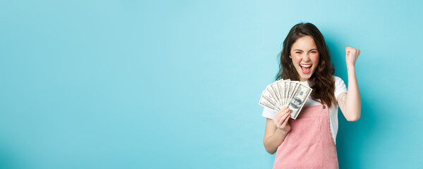 Wall Mural - Lucky young woman looks excited, shouting from satisfaction and triumph, winning money, holding dollar bills and making fist pump, standing over blue background