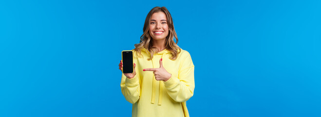 Proud cheerful young blond woman advertise new mobile game or application, look camera and smiling white teeth as pointing smartphone display, recommend download or try out