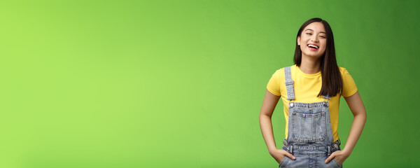 Wall Mural - Carefree enthsuastic outgoing attractive teenage asian girl having fun laughing friends, hold hands overalls pockets smiling toothy, take party easygoing conversation, stay positive green background