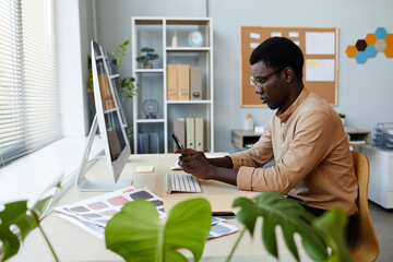 Wall Mural - Side view portrait of young black man using smartphone at workplace in minimal office setting with plants