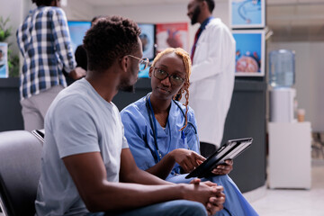 African american nurse and patient talking about disease, doing checkup consultation with sick man in hospital waiting area. Medical assistant examining person to give medication, health center.