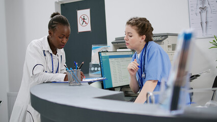 Diverse medical team working at hospital reception counter to help patients with consultation appointments and checkup visits. Doctor and nurse checking report papers. Handheld shot.