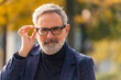 © PoppyPix - Eyeglasses commercial concept. Outdoor closeup shot of handsome elegant caucasian man in his 60s with gray facial hair touching his black-frame glasses with one hand. Warm autumn sun shining on part