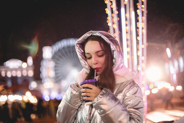 Happy young woman drinking mulled wine while standing in city during Christmas holidays at night