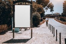 Mockup Of An Empty Vertical Ad Poster In Urban Settings On The Pavement Next To A Cobbled Road; A Blank Street Banner Template; An Outdoor Billboard Placeholder Mock-up On The Sunny Street