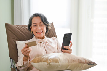 Wall Mural - Senior Asian female using mobile banking application while relaxing in her living room.