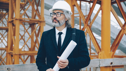 Wall Mural - Portrait of serious mature man architect standing in construction site with blueprint looking around wearing white helmet and suit. People and occupation concept.