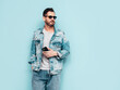 © halayalex - Handsome confident stylish hipster lambersexual model. Sexy man dressed in jeans jacket. Fashion male isolated on blue wall in studio. In sunglasses. Holding smartphone. Looking at phone screen, using
